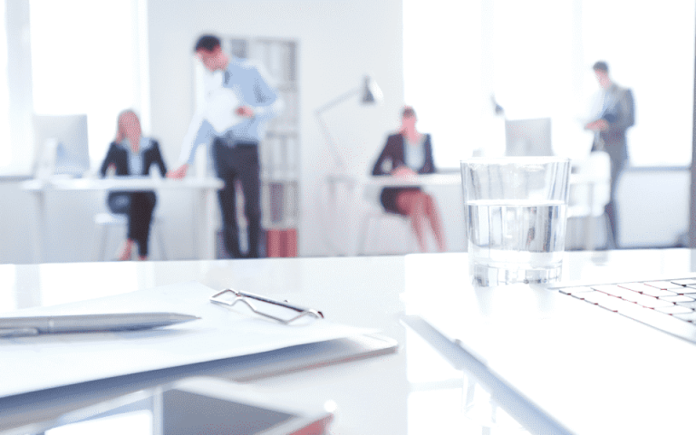 A focused shot of a work desk with a laptop, water cup and notepad with people in the blurred in the background